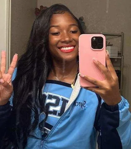Syrai Smith smiling and holding up a peace sign, wearing her light blue cheer jacket.