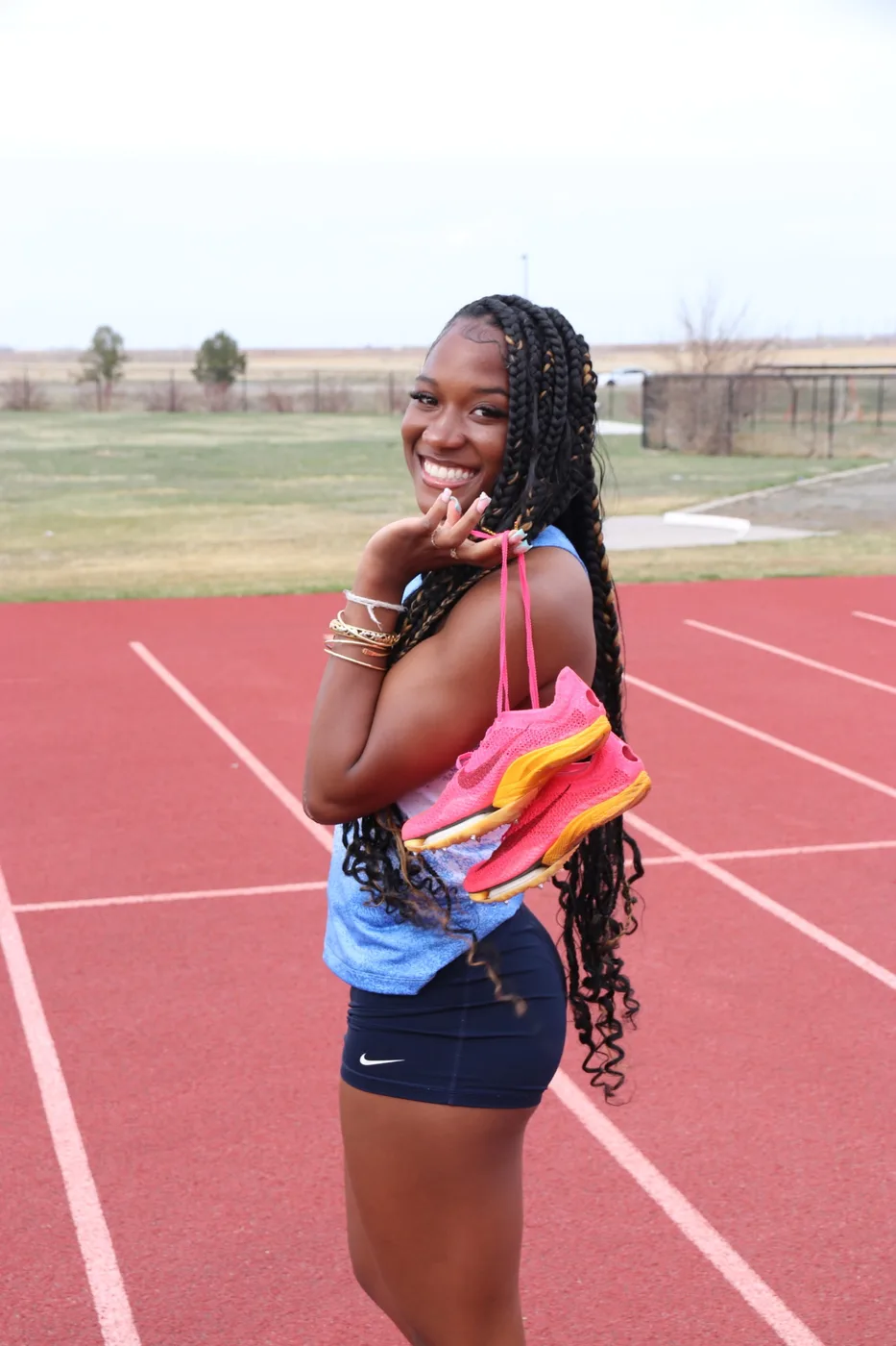 Syrai Smith smiling on a running track, holding a pair of pink track spikes over her shoulder by the laces.
